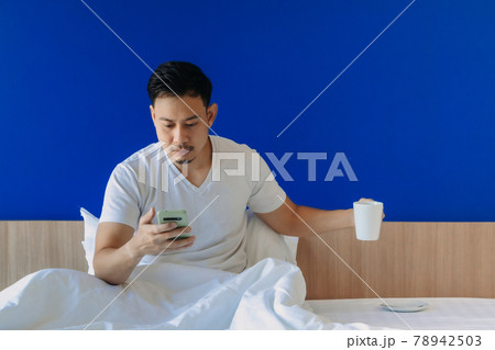 Man with smartphone and coffee on the bed on blue copy space background. 78942503