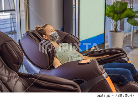 Beautiful young woman wearing a medical mask due to the coronavirus COVID 19 relaxing on the massage chair in airport or in the mall Beautiful young woman wearing a medical mask due to the coronavirus COVID 19 relaxing on the massage chair in airport or in the mall 78950647