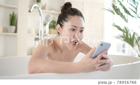 Excited young multi ethnic asian woman celebrating bid win success victory or getting ecommerce shopping offer on smartphone, during morning daily body care and beauty procedure in bathroom Excited young multi ethnic asian woman celebrating bid win success victory or getting ecommerce shopping offer on smartphone, during morning daily body care and beauty procedure in bathroom 78956766