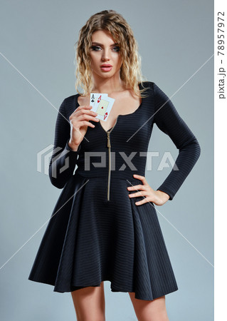Blonde woman in black short dress showing two playing cards, posing against gray background. Gambling entertainment, poker, casino. Close-up. Blonde woman in black short dress showing two playing cards, posing against gray background. Gambling entertainment, poker, casino. Close-up. 78957972