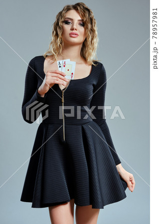Blonde maiden with curly hair, in black short dress showing two aces, posing against gray background. Gambling entertainment, poker, casino. Close-up. Blonde maiden with curly hair, in black short dress showing two aces, posing against gray background. Gambling entertainment, poker, casino. Close-up. 78957981