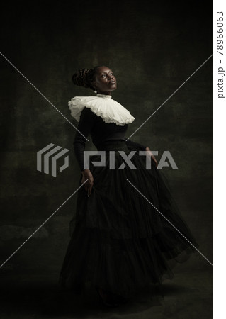 Medieval African young woman in black vintage dress with big white collar posing isolated on dark green background. Concept of comparison of eras, modernity and renaissance. Medieval African young woman in black vintage dress with big white collar posing isolated on dark green background. Concept of comparison of eras, modernity and renaissance. 78966063