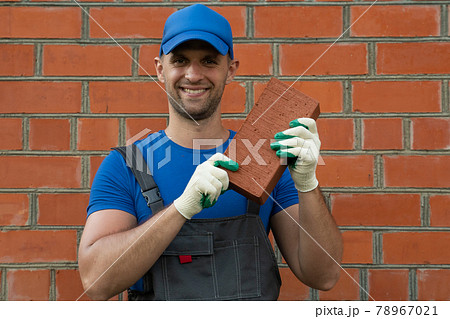 A man wearing a cap and gloves shows a brick to the camera in a construction concept A man wearing a cap and gloves shows a brick to the camera in a construction concept 78967021