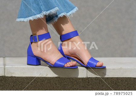 Women's legs in blue denim jeans and sandals in the city street. Trendy elegant casual outfit. Details of everyday summer look. 78975025
