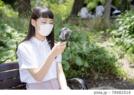 公園のベンチに座り、マスクをして手持ち扇風機で涼む少女 78982019