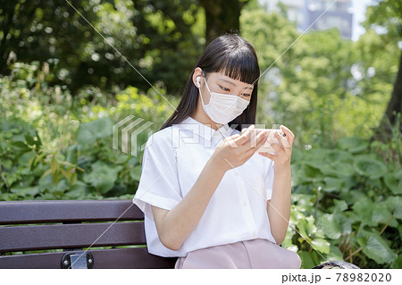 公園のベンチに座り、マスクをしてスマホで動画を観る少女 78982020