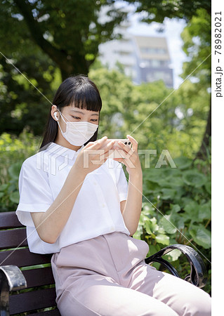 公園のベンチに座り、マスクをしてスマホで動画を観る少女 78982021