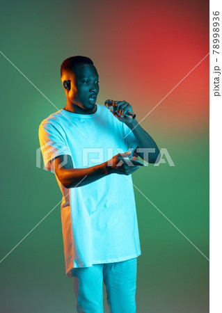 Young african man speaking with microphone on neon gradient background. Concept of emotion, action, facial expression. Young african man speaking with microphone on neon gradient background. Concept of emotion, action, facial expression. 78998936