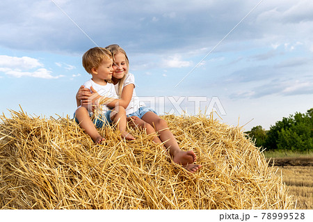 Pair of two cute adorable little caucasian sibling kids enjoy sitting on haystack bale and hug together at country nature farm on warm summer day. Brother and sister friendship, love and care concept Pair of two cute adorable little caucasian sibling kids enjoy sitting on haystack bale and hug together at country nature farm on warm summer day. Brother and sister friendship, love and care concept 78999528