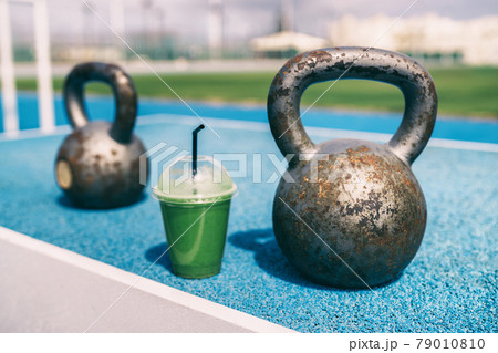Health and fitness green smoothie detox drink at gym with kettlebells weights at outdoor training fitness center. Plastic cup of vegetable juice morning breakfast next to kettlebell weight equipment Health and fitness green smoothie detox drink at gym with kettlebells weights at outdoor training fitness center. Plastic cup of vegetable juice morning breakfast next to kettlebell weight equipment 79010810