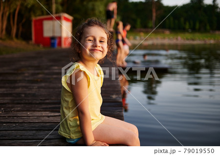 Happy girl on a pier smiles looking at camera. Summer leisure activities concepts. Heat summer, summer evenings in nature, spending time in the fresh air Happy girl on a pier smiles looking at camera. Summer leisure activities concepts. Heat summer, summer evenings in nature, spending time in the fresh air 79019550