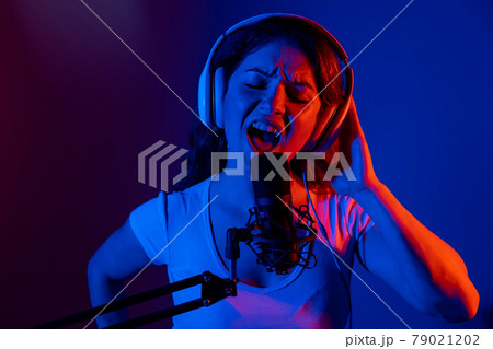 Caucasian woman in headphones singing into a microphone in neon light on a black background. An emotional girl is recording a song in a recording studio 79021202