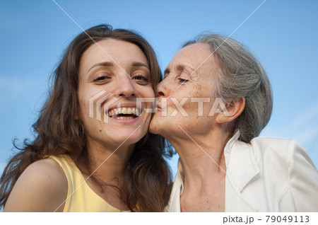 Senior mother with gray hair with her adult daughter looking at the camera in the garden and hugging each other during sunny day outdoors Senior mother with gray hair with her adult daughter looking at the camera in the garden and hugging each other during sunny day outdoors 79049113