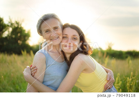 Senior mother with gray hair with her adult daughter looking at the camera in the garden and hugging each other during sunny day outdoors Senior mother with gray hair with her adult daughter looking at the camera in the garden and hugging each other during sunny day outdoors 79049118