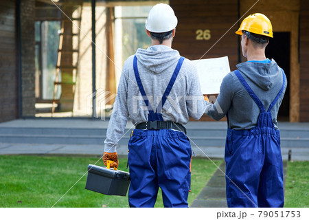 Plan it. Rear view of two builders standing outdoors with an open blueprint discussing the interior of a half completed cottage house 79051753