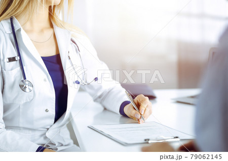 Unknown woman- doctor is listening to her patient, while sitting together at the desk in the sunny cabinet in a clinic. Female physician with a stethoscope is writing at clipboard, close up. Perfect Unknown woman- doctor is listening to her patient, while sitting together at the desk in the sunny cabinet in a clinic. Female physician with a stethoscope is writing at clipboard, close up. Perfect 79062145