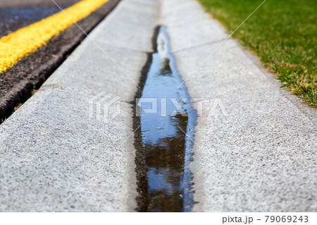 flowing rainwater in the canal is a cement ditch of a drainage system on the side of wet asphalt road with yellow markings and a green lawn on roadside. flowing rainwater in the canal is a cement ditch of a drainage system on the side of wet asphalt road with yellow markings and a green lawn on roadside. 79069243