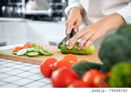 Close up of asian housewife wearing apron and using knife to slice cucumber and tomato on chopping board 79088782