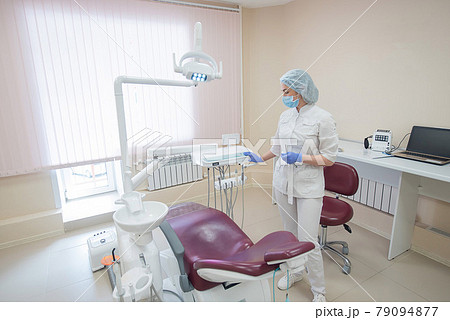 Girl dentist, in white clothes, mask and cap stands next to a modern dental chair in a large, simple, medical office Girl dentist, in white clothes, mask and cap stands next to a modern dental chair in a large, simple, medical office 79094877