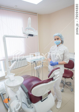 Girl dentist, in white clothes, mask and cap stands next to a modern dental chair in a large, simple, medical office 79094880