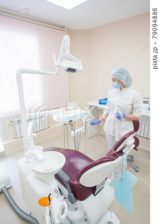 Girl dentist, in white clothes, mask and cap stands next to a modern dental chair in a large, simple, medical office 79094886