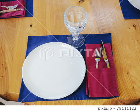 White plate with a crystal glass fork and knife on a blue napkin on a wooden light table. New shiny cutlery. Closeup photo White plate with a crystal glass fork and knife on a blue napkin on a wooden light table. New shiny cutlery. Closeup photo 79111122