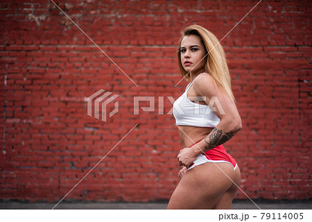 Young woman bodybuilder posing while standing with her back against a brick wall. Outdoor sports in isolation. A strong and muscular girl is engaged in fitness on a sports field. 79114005