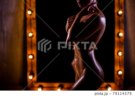 A young, naked, Caucasian girl with a very slender figure poses against a large mirror with lights in the loft style. The skin is oiled and shiny. Sexy tan. 79114379