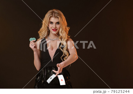 Blonde girl in black dress and necklace. Smiling, showing two green chips, throwing something, posing on brown background. Poker, casino. Close-up Blonde girl in black dress and necklace. Smiling, showing two green chips, throwing something, posing on brown background. Poker, casino. Close-up 79125244