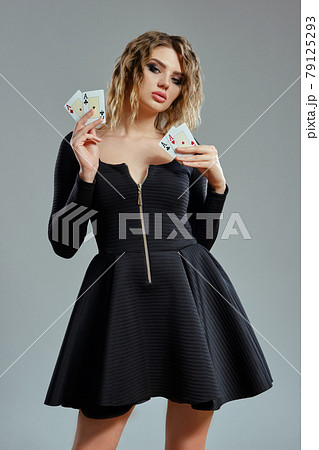 Blonde lady in black short dress showing four playing cards, posing against gray background. Gambling entertainment, poker, casino. Close-up. Blonde lady in black short dress showing four playing cards, posing against gray background. Gambling entertainment, poker, casino. Close-up. 79125293