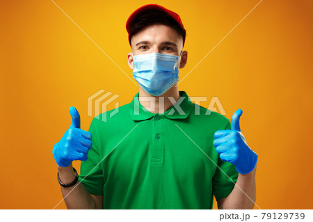 Young delivery man in green t-shirt and cap wearing face mask against yellow background 79129739