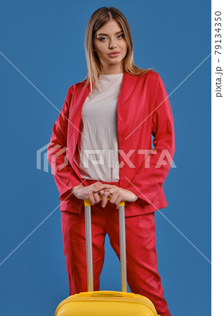 Alluring blonde girl in red pantsuit and white blouse. She is looking at you, holding yellow suitcase, posing on blue background. Close-up Alluring blonde girl in red pantsuit and white blouse. She is looking at you, holding yellow suitcase, posing on blue background. Close-up 79134350