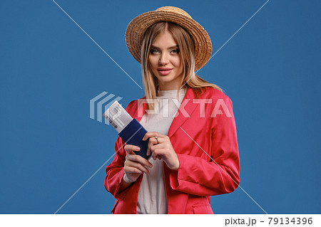 Blonde female in straw hat, white blouse and red pantsuit. She smiling, holding passport and ticket while posing on blue studio background. Close-up Blonde female in straw hat, white blouse and red pantsuit. She smiling, holding passport and ticket while posing on blue studio background. Close-up 79134396