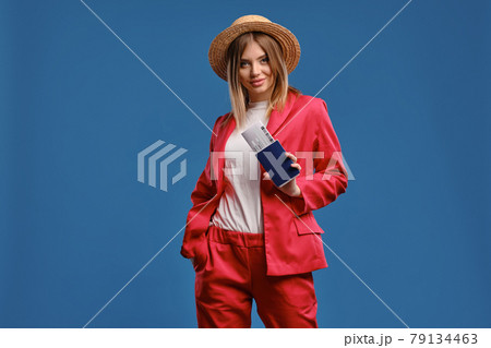 Blonde model in straw hat, white blouse and red pantsuit. She smiling, showing passport and ticket while posing on blue studio background. Close-up Blonde model in straw hat, white blouse and red pantsuit. She smiling, showing passport and ticket while posing on blue studio background. Close-up 79134463