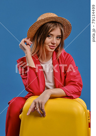 Blonde girl in straw hat, white blouse, red pantsuit. She sitting leaning on yellow suitcase, posing on blue background. Close-up, copy space 79134499