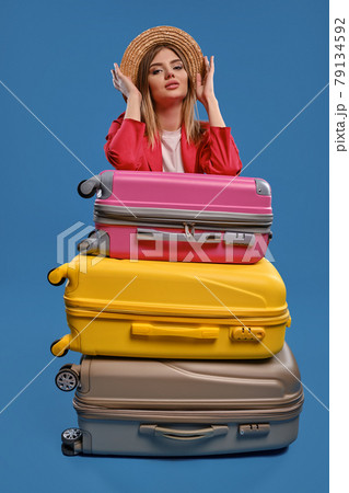 Blonde female in straw hat, white blouse, red jacket. Sitting leaning on colorful suitcases, posing on blue background. Travelling concept. Close-up 79134592