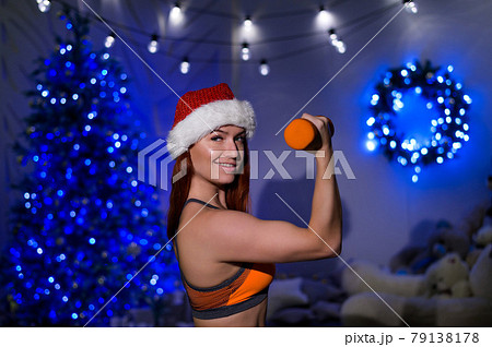 Beautiful red-haired female athlete in a Santa Claus hat posing in a dark studio. Fitness woman in sportswear holds a dumbbell in her hands on the background of the New Year tree. Beautiful red-haired female athlete in a Santa Claus hat posing in a dark studio. Fitness woman in sportswear holds a dumbbell in her hands on the background of the New Year tree. 79138178