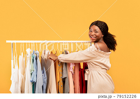 Seasonal shopping concept. Black female choosing clothes from rack, following latest fashion trends on orange background Seasonal shopping concept. Black female choosing clothes from rack, following latest fashion trends on orange background 79152226