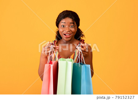 Summer shopping concept. Surprised black woman with paper bags looking at camera on orange studio background Summer shopping concept. Surprised black woman with paper bags looking at camera on orange studio background 79152327