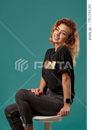 Ginger girl with a curly hair, wearing black t-shirt with a golden stripe is posing sitting on a chair against blue studio background. Close-up shot. Ginger girl with a curly hair, wearing black t-shirt with a golden stripe is posing sitting on a chair against blue studio background. Close-up shot. 79154130