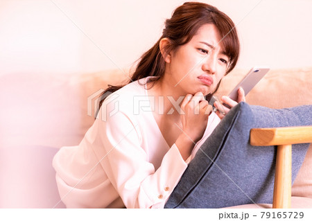 リビングのソファに寝そべってスマホの画面を見る不機嫌な女性 79165729