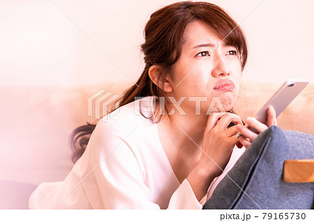 リビングのソファに寝そべってスマホの画面を見る不機嫌な女性 79165730