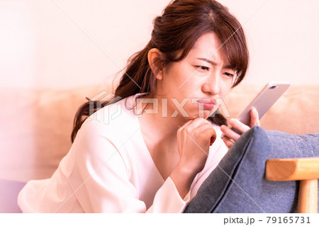 リビングのソファに寝そべってスマホの画面を見る不機嫌な女性 79165731