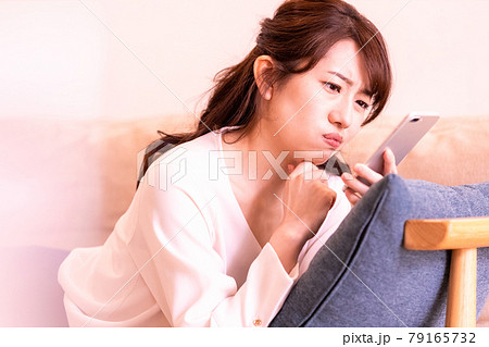 リビングのソファに寝そべってスマホの画面を見る不機嫌な女性 79165732