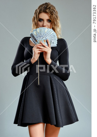 Blonde girl in black short dress is holding some cash, posing against gray background. Gambling entertainment, poker, casino. Close-up. Blonde girl in black short dress is holding some cash, posing against gray background. Gambling entertainment, poker, casino. Close-up. 79173362