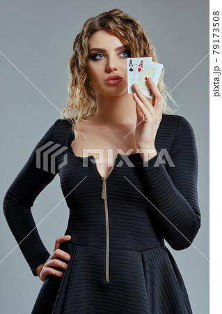 Blonde woman in black dress showing two playing cards, posing against gray background. Gambling entertainment, poker, casino. Close-up. 79173508