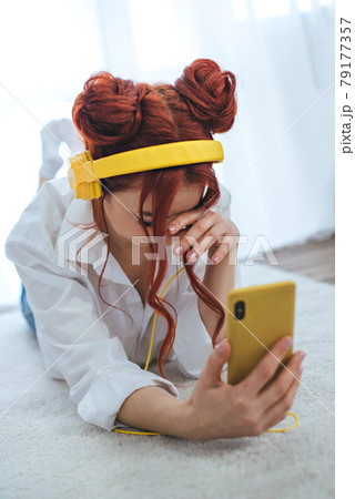 A girl sits on the bed, looks out the window and listens to music in yellow headphones. 79177357