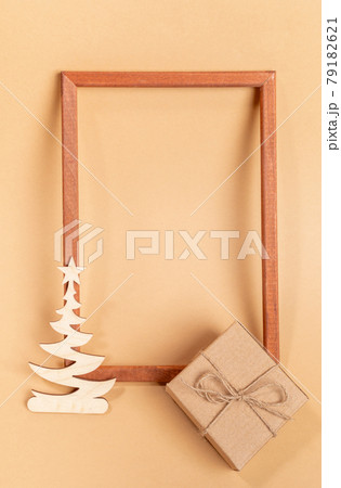Festive minimal layout with empty wooden frame, small wooden carved decoration - Xmas tree, cardboard gift box on beige background. Christmas or New Year mock up. Top view. Copy space. Vertical photo. Festive minimal layout with empty wooden frame, small wooden carved decoration - Xmas tree, cardboard gift box on beige background. Christmas or New Year mock up. Top view. Copy space. Vertical photo. 79182621