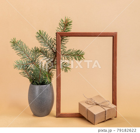 Festive trendy minimal composition with fir tree branches in grey stone vase, empty wooden frame and cardboard gift box on beige background. Christmas or New Year mock up. Square photo. 79182622