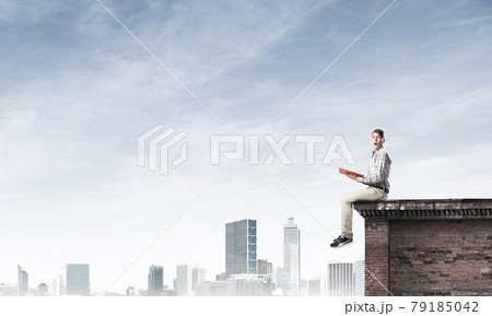 Man on roof edge reading book and cityscape at background 79185042